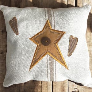 May include: Square white pillow with a large gold star and a brown button in the center. Two brown heart shapes are on the sides. The pillow has a striped design down the center.