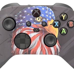 May include: A black Xbox controller with a custom design featuring an American bald eagle with an American flag design. The eagle's head is facing the right side of the controller. The controller has a black d-pad, black buttons, and black triggers.