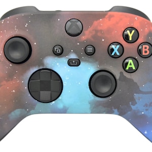 May include: A black Xbox controller with a galaxy-themed design. The controller features a blue, red, and black nebula pattern with white stars. The controller has a black D-pad, black face buttons, and black triggers.