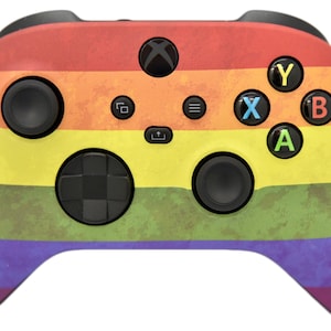 May include: A black Xbox controller with a rainbow-colored design. The controller has a textured surface and features the standard Xbox buttons: A, B, X, Y, and a directional pad.