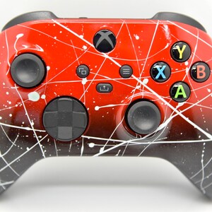 May include: A red and black Xbox controller with a white splatter paint design. The controller has a black d-pad, black buttons, and black triggers.