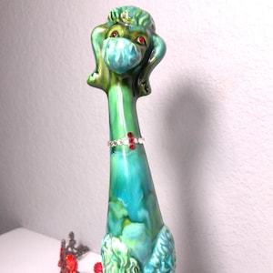 May include: A ceramic poodle figurine in shades of green and teal, adorned with a red jeweled collar. The poodle has a long neck and is seated. A red jeweled crown is in the background.