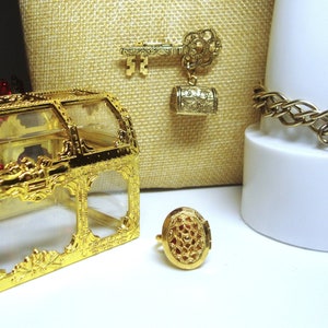 May include: A gold-colored treasure chest with a clear lid, a gold-colored key charm with a barrel pendant, a gold-colored charm bracelet with a barrel pendant, and a gold-colored ring with an ornate design.