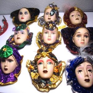 May include: Nine porcelain masquerade masks with colorful fabric and feather embellishments. The masks feature different facial expressions and are decorated with gold, green, blue, red, and purple accents.
