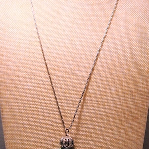 May include: A silver chain necklace with a green malachite pendant shaped like a bottle. The bottle has a silver cap with an ornate design.