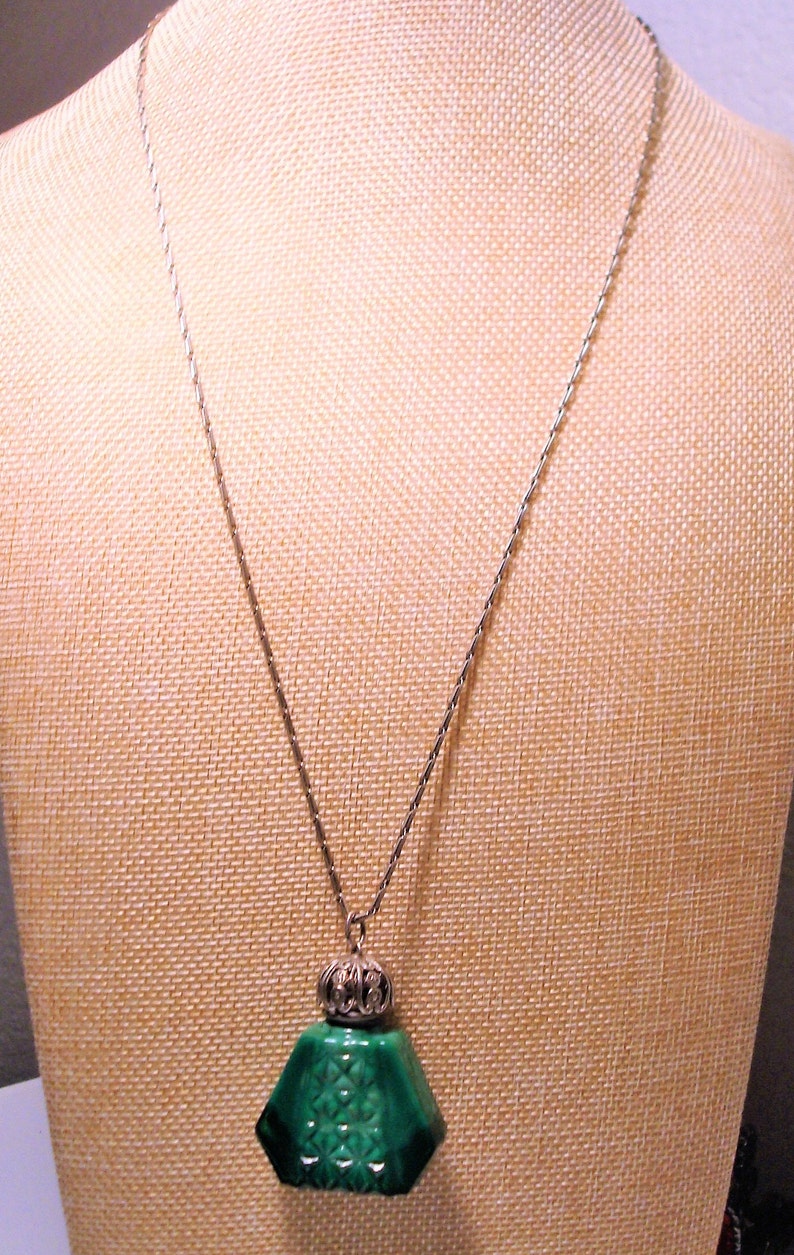 May include: A silver chain necklace with a green malachite pendant shaped like a bottle. The bottle has a silver cap with an ornate design.