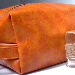 Pu&ograve; includere: Un beauty case in pelle arancione con chiusura a zip e manico. Accanto alla borsa c'&egrave; una bottiglia di profumo in vetro trasparente con tappo dorato e dettagli decorativi. La borsa e la bottiglia sono su una superficie bianca.