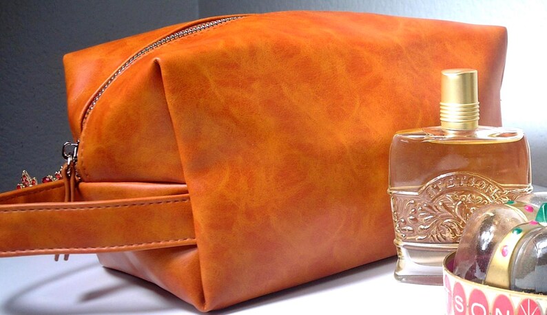 Pu&ograve; includere: Un beauty case in pelle arancione con chiusura a zip e manico. Accanto alla borsa c'&egrave; una bottiglia di profumo in vetro trasparente con tappo dorato e dettagli decorativi. La borsa e la bottiglia sono su una superficie bianca.