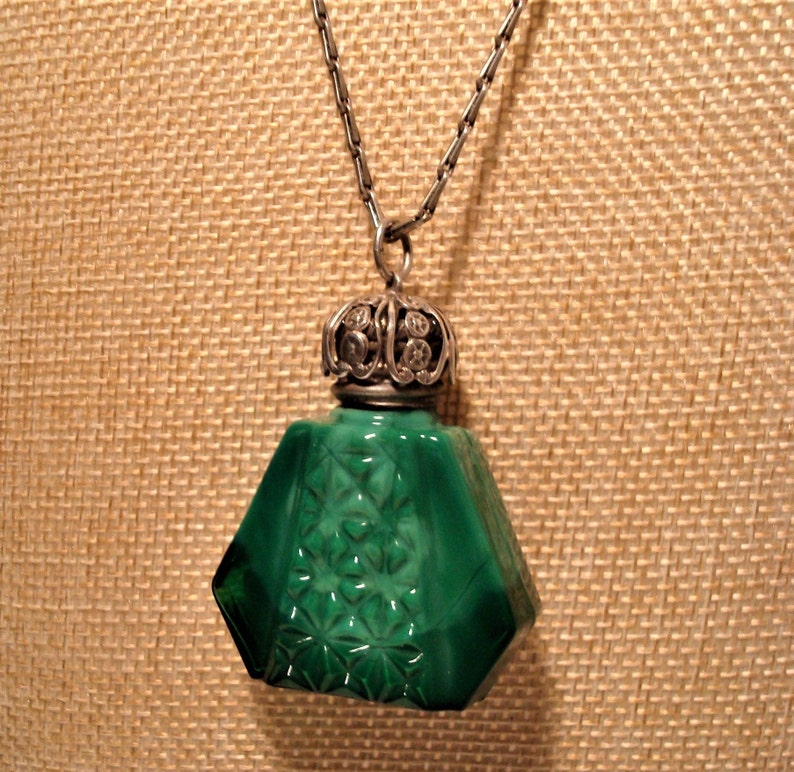May include: A green glass bottle pendant with a silver filigree top and a silver chain. The bottle has a faceted design and is reminiscent of a vintage perfume bottle.
