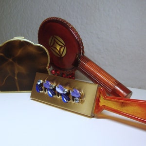 May include: A set of vintage vanity items including a compact with a brown marbled design, a red hand mirror with a gold design, and a gold and orange brush with blue and purple rhinestones.