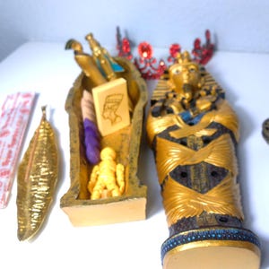 May include: A collection of Egyptian-themed decorative items. Includes a golden sarcophagus, a pharaoh figurine, a small incense burner, a golden leaf, and a red jeweled crown. The sarcophagus is open, revealing a small figure and other items.