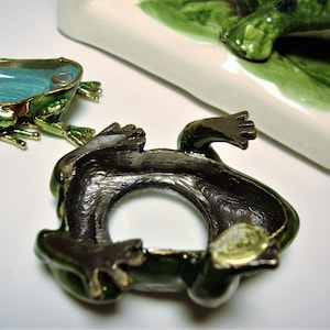 May include: A green frog figurine with a hollow interior, the frog is made of metal and has a gold trim. The frog is lying on its back with its legs in the air. The frog is a trinket box.