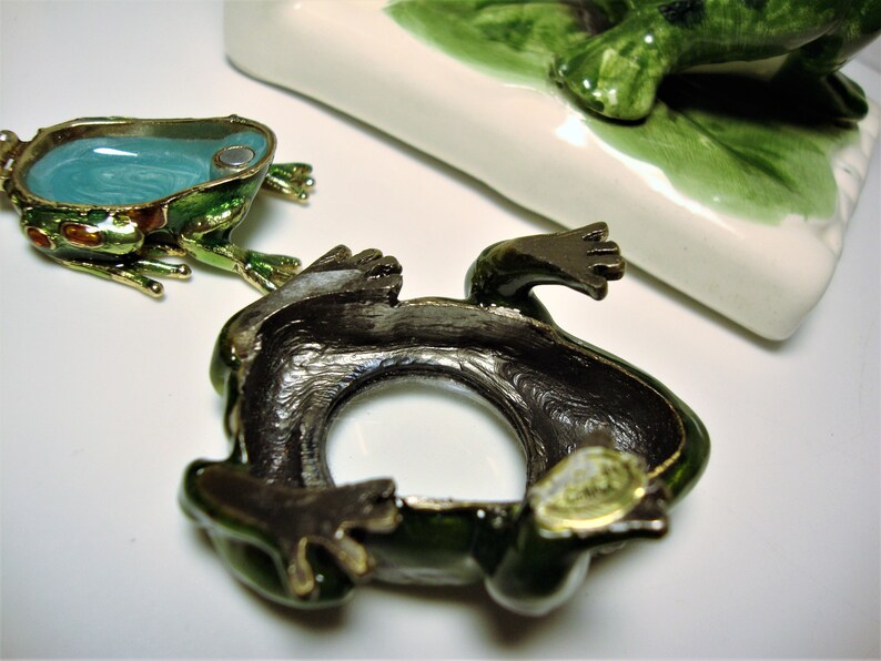 May include: A green frog figurine with a hollow interior, the frog is made of metal and has a gold trim. The frog is lying on its back with its legs in the air. The frog is a trinket box.