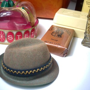 Pu&ograve; includere: Un cappello fedora grigio con tesa blu scuro e rifiniture a zig-zag gialle. Accanto al cappello ci sono una saponetta, un contenitore decorativo e un flacone di profumo.