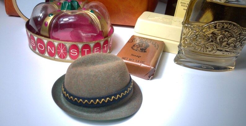 Pu&ograve; includere: Un cappello fedora grigio con tesa blu scuro e rifiniture a zig-zag gialle. Accanto al cappello ci sono una saponetta, un contenitore decorativo e un flacone di profumo.