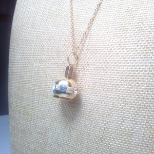 May include: A gold-colored pendant necklace featuring a small, round charm with blue and clear crystal accents. The charm has a cylindrical top and hangs from a delicate gold chain. The necklace is displayed on a textured, off-white surface.