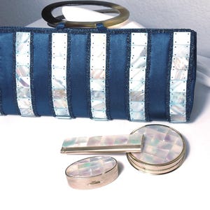 May include: A navy blue clutch with a clear handle, and three matching accessories. The clutch is rectangular with vertical white and mother-of-pearl accents. The accessories include a rectangular case, a round compact, and an oval compact.