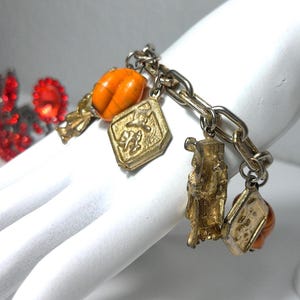 May include: A vintage charm bracelet with gold-toned charms and a silver-toned chain. The charms include an orange bead, a gold-toned figure, and two gold-toned pendants. The bracelet is displayed on a white surface.
