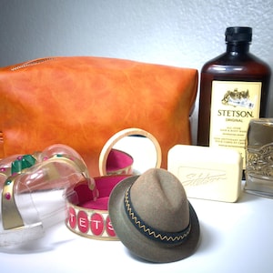 Pu&ograve; includere: Un assortimento di articoli per la cura della persona e accessori vintage. Include una borsa da toilette in pelle marrone, un piccolo cappello fedora, una bottiglia di colonia Stetson, una saponetta e una corona decorativa.