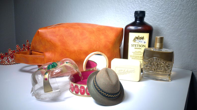 Pu&ograve; includere: Un assortimento di articoli per la cura della persona e accessori vintage. Include una borsa da toilette in pelle marrone, un piccolo cappello fedora, una bottiglia di colonia Stetson, una saponetta e una corona decorativa.