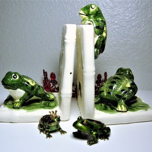 May include: A set of ceramic frog bookends with a green and white design. The bookends are shaped like frogs sitting on a green leaf. There are two smaller green frog figurines in the foreground.
