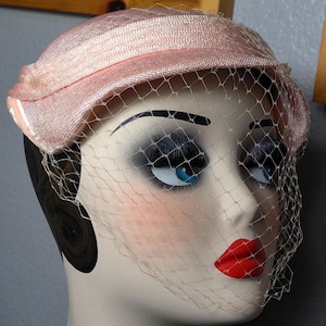 May include: A vintage-style pink hat with a matching veil, displayed on a mannequin head. The hat has a structured design with a slightly curved brim. The veil is a fine mesh, adding a touch of elegance. The mannequin has blue eyes and red lipstick.