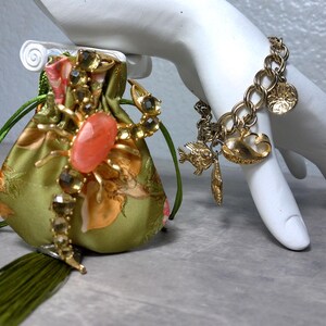 May include: A small olive green satin drawstring pouch featuring a large gold-tone scorpion brooch with a central oval coral-colored stone and a long green tassel, paired with a gold-tone charm bracelet featuring various sea-themed metal charms.