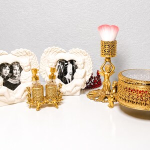May include: A collection of decorative items including two cream-colored heart-shaped frames with cherub motifs, a gold-tone perfume bottle set, a gold-tone brush holder with a pink-tipped brush, a red crown, and a round gold-tone jewelry box.