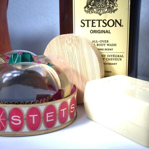 Pu&ograve; includere: Una collezione di prodotti per la cura della persona tra cui una saponetta, una bottiglia di Stetson Original All-Over Body Wash e un contenitore decorativo con la scritta "SONSTETS". Il contenitore ha un bordo dorato e dettagli rossi e bianchi.
