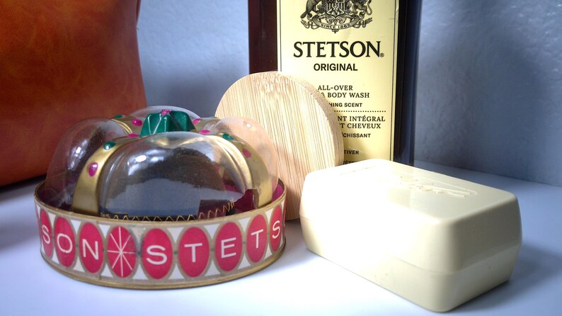 Pu&ograve; includere: Una collezione di prodotti per la cura della persona tra cui una saponetta, una bottiglia di Stetson Original All-Over Body Wash e un contenitore decorativo con la scritta "SONSTETS". Il contenitore ha un bordo dorato e dettagli rossi e bianchi.