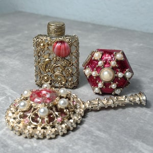May include: An ornate perfume bottle, a hexagonal container, and a hand mirror, all with gold filigree and red accents. The pieces are embellished with faux pearls. The bottle has a decorative stopper.