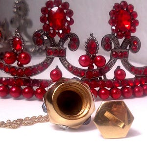 May include: Close-up of a red and silver tiara with large red gemstones and smaller red beads. A gold-colored bottle stopper and a gold-colored, hexagonal object are in the foreground. A gold chain is also visible.