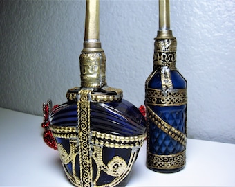 May include: Two blue glass bottles with gold metal accents and intricate designs. The bottles have a decorative, ornate look.