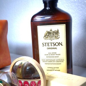 Pu&ograve; includere: Una bottiglia marrone di Stetson Original All-Over Hair & Body Wash con tappo nero. Sono visibili anche un piccolo specchio rotondo, una saponetta e un contenitore con la scritta "Stetson". L'etichetta recita "Citrus & Vetiver."