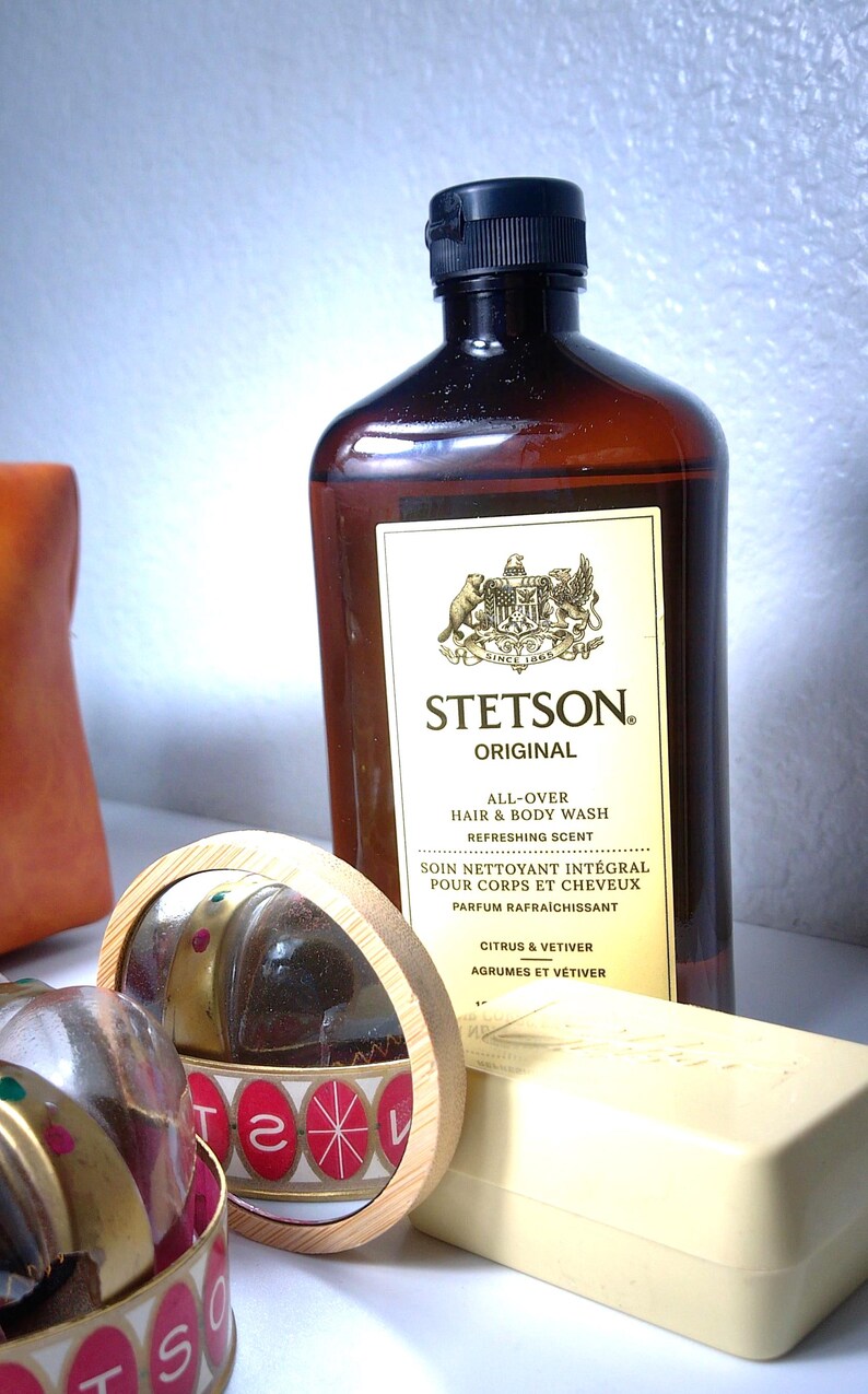 Pu&ograve; includere: Una bottiglia marrone di Stetson Original All-Over Hair & Body Wash con tappo nero. Sono visibili anche un piccolo specchio rotondo, una saponetta e un contenitore con la scritta "Stetson". L'etichetta recita "Citrus & Vetiver."