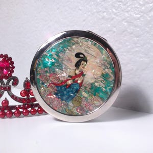 May include: A silver compact mirror with an Asian-inspired design. The mirror features a woman in a flowing gown, surrounded by flowers and clouds. A red jeweled crown is in the background.