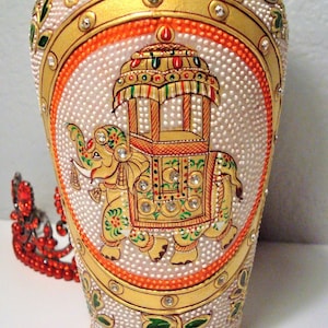 May include: A white ceramic vase with gold and orange accents. The vase is decorated with an elephant carrying a palanquin, all painted in intricate detail. The vase is adorned with small, sparkling rhinestones.