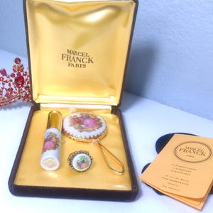 May include: A vintage Marcel Franck Paris vanity set in a velvet-lined box. The set includes a perfume bottle, compact mirror, and ring, all with painted scenes. A red jeweled tiara is to the left, and a product information card is on the right.
