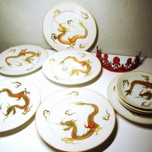 May include: Set of six white ceramic plates with gold trim and a red dragon design. The plates are arranged in a circle with a red crown in the center.