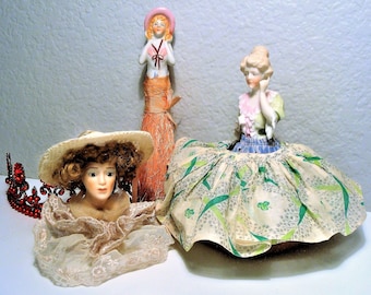 May include: Three porcelain dolls with painted faces and detailed clothing. The doll on the left is a bust wearing a white hat and a lace dress. The doll in the center is wearing a green and white dress with a floral pattern. The doll on the right is wearing a pink hat and a white dress with a floral pattern.