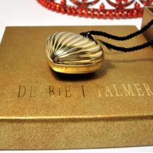 May include: A gold heart-shaped locket with a ribbed design, on a black cord necklace, in a gold box with the text "DE BEIJ PALMER TM".