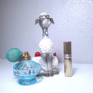 May include: A collection of vintage decorative items, including a gray and white poodle figurine, a blue glass perfume bottle with a teal atomizer, and a gold-toned perfume bottle. The poodle has a green gem collar.
