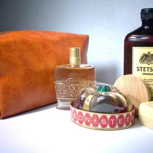 Pu&ograve; includere: Un beauty case in pelle arancione, una bottiglia di colonia, una bottiglia di Stetson Original All-Over Body Wash, una saponetta e un contenitore decorativo con la scritta "STETSON". Gli articoli sono disposti su una superficie bianca.