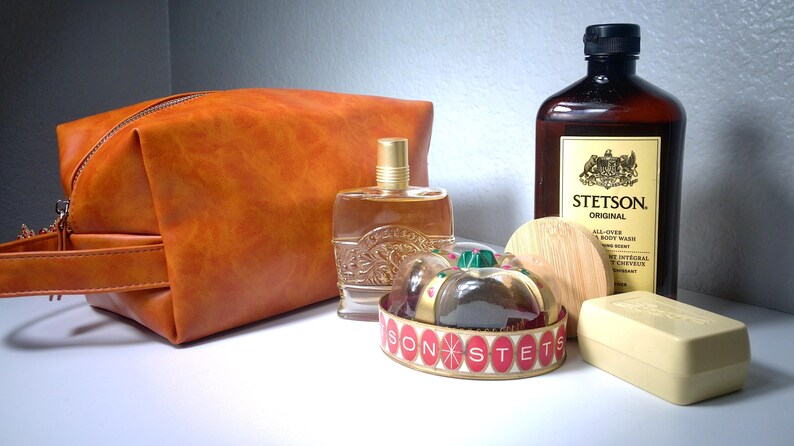 Pu&ograve; includere: Un beauty case in pelle arancione, una bottiglia di colonia, una bottiglia di Stetson Original All-Over Body Wash, una saponetta e un contenitore decorativo con la scritta "STETSON". Gli articoli sono disposti su una superficie bianca.
