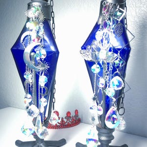 May include: Two decorative blue glass bottles with silver accents and hanging crystal ornaments. Each bottle has a silver cap and chain. One bottle features a crescent moon and star design. The bottles are on a black metal stand.