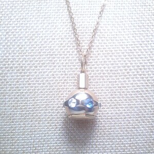 May include: A silver-toned pendant necklace featuring a small, rounded charm with two blue gemstones. The charm has a geometric design and is suspended from a delicate chain. The background is a textured, off-white fabric.