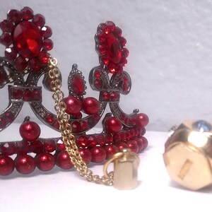May include: A decorative crown with red jewels and a gold chain, next to a gold and jewel-encrusted sphere. The crown features a dark metal frame and is adorned with red gems and pearls. The sphere has a geometric design.