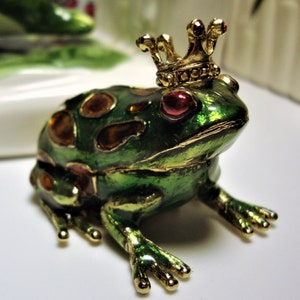May include: A green frog figurine with gold accents and a jeweled crown. The frog has red eyes and is decorated with gold and orange jewels.
