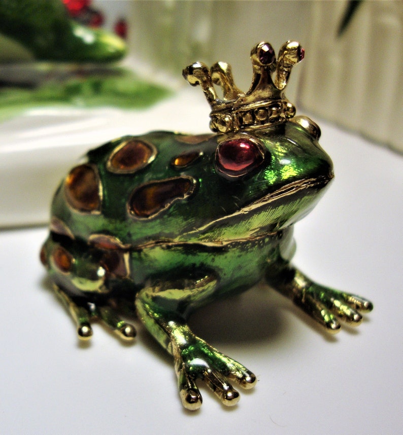 May include: A green frog figurine with gold accents and a jeweled crown. The frog has red eyes and is decorated with gold and orange jewels.