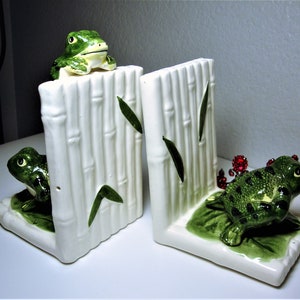 May include: A pair of ceramic bookends featuring green frogs on a white bamboo design. One frog is perched on top of the bamboo, while the other is sitting on a green leaf.